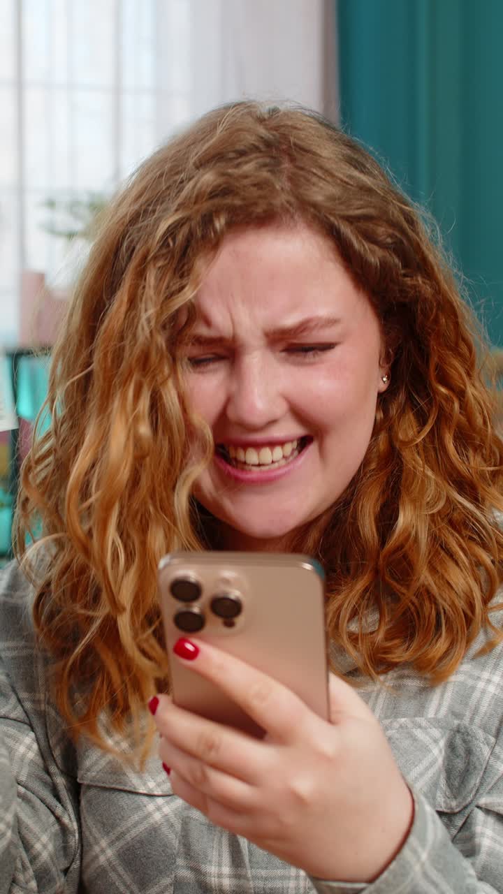 Young woman sits on sofa smiling happily showing thumbs up after receiving money sms on smartphone