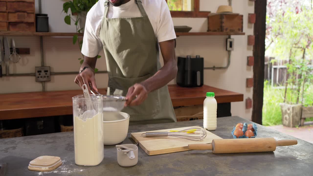 Man in apron in kitchen making dry-mix scooping flour from jar into sieve whisking on-camera