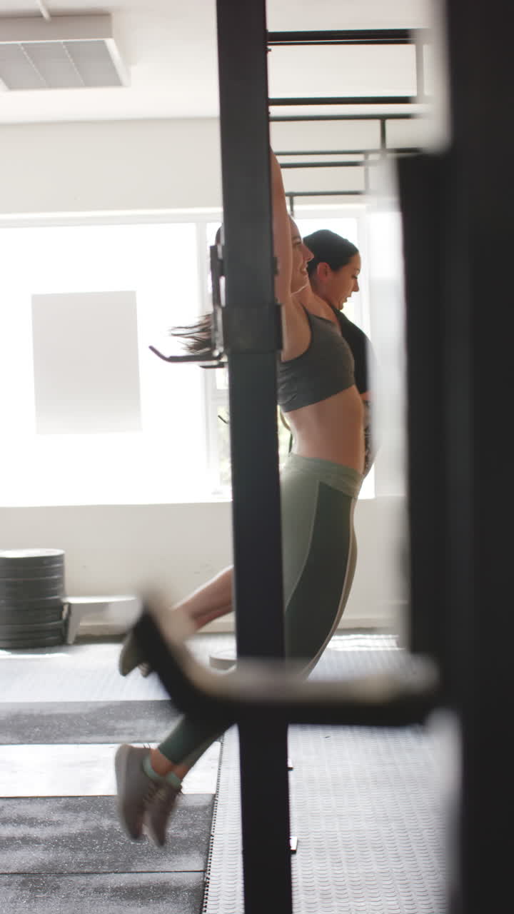 Vertical video of two unaltered diverse women doing pull ups at gym in slow motion