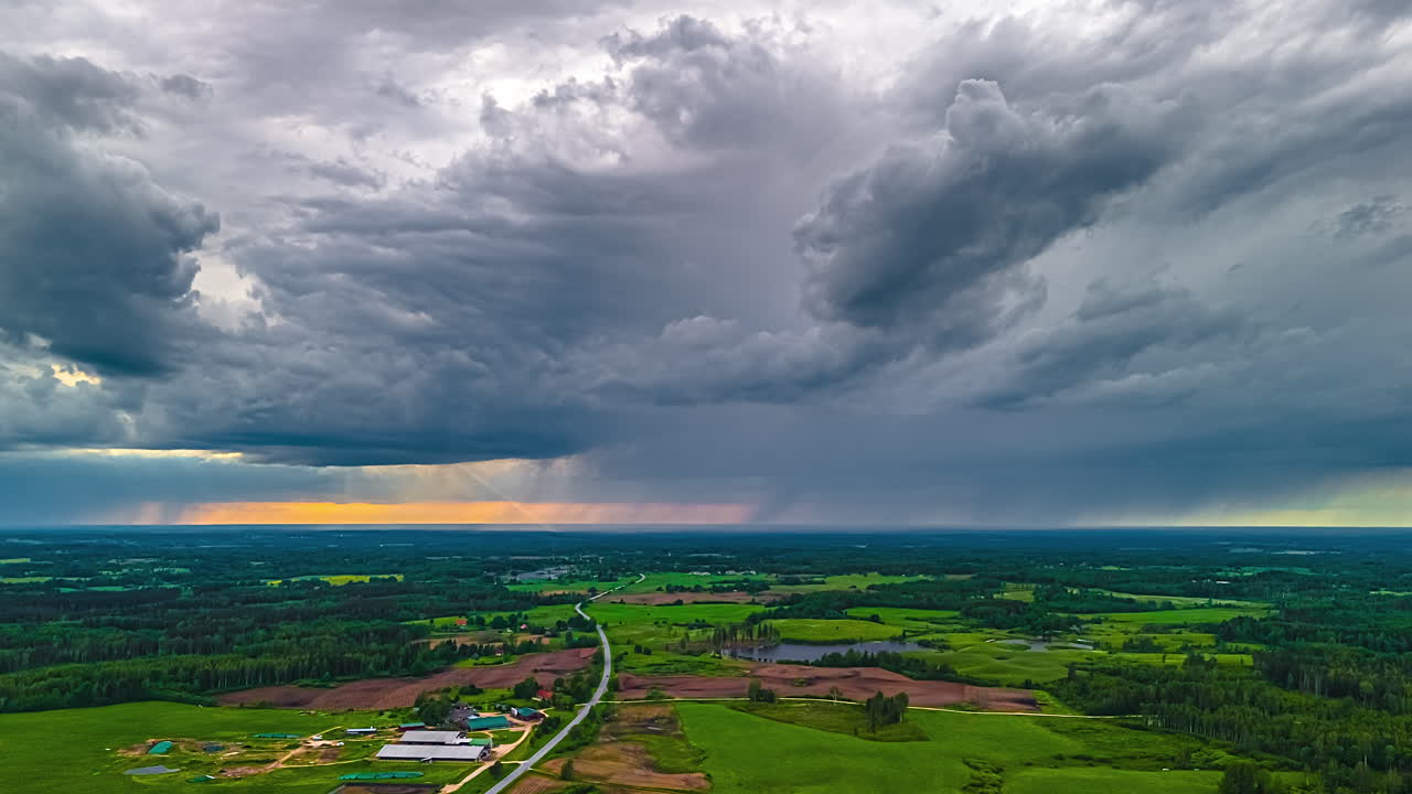 Dramatic timelapse of dark storm clouds rolling across rural green landscape with orange glow on horizon, static