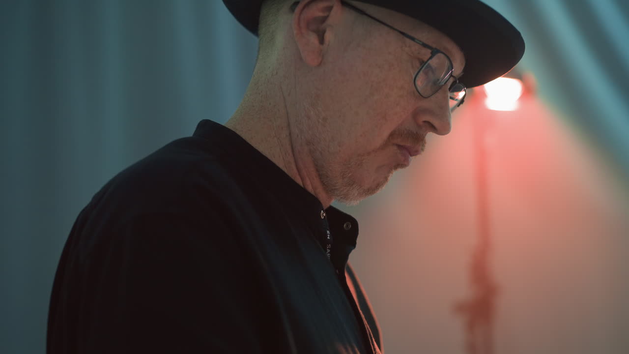 Elderly man wearing a black shirt and hat, reflecting pensively, with glasses, captured in profile against a softly blurred multicolored light background, emphasizing a deep, contemplative mood