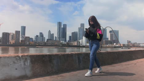 full shot of a young girl writing on the mobile phone near the river with a city in the background