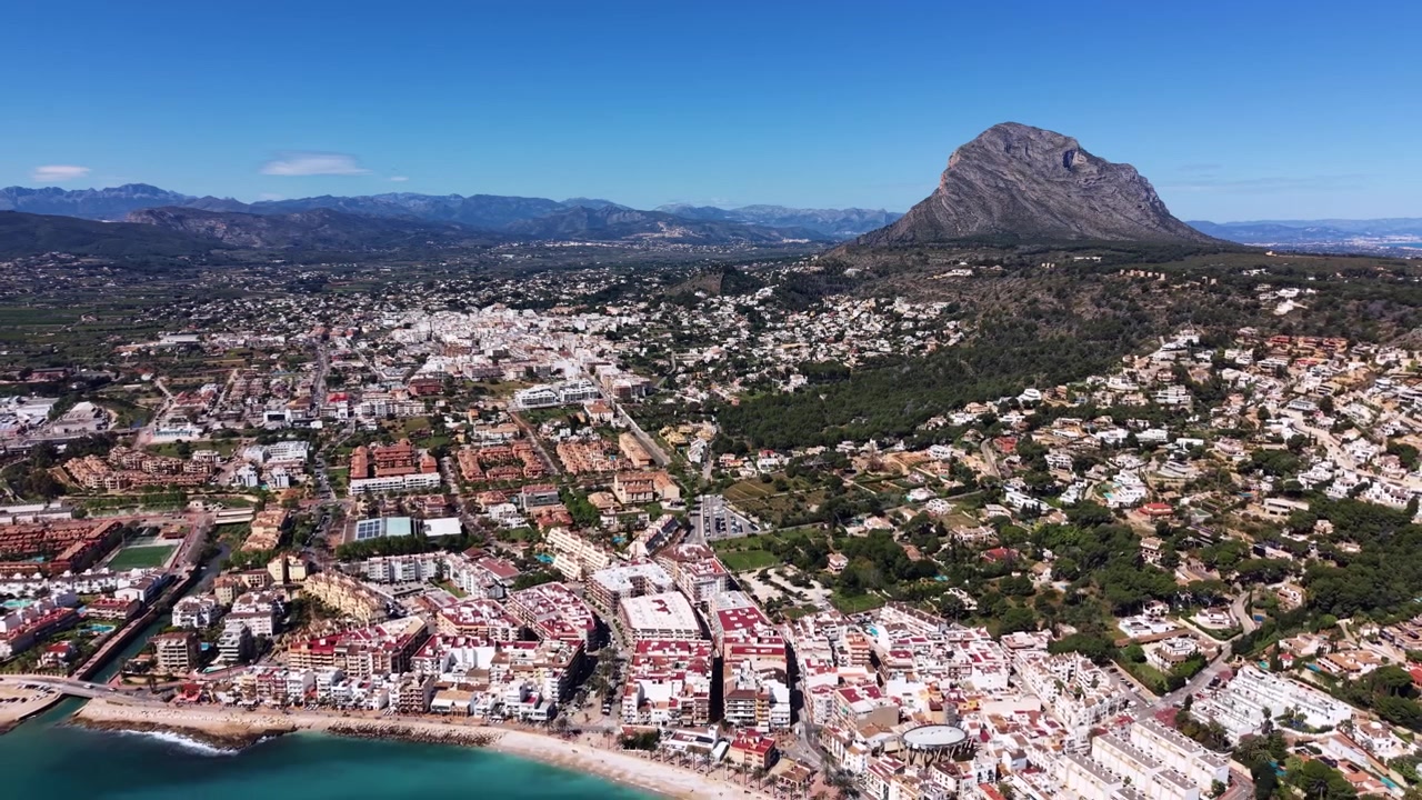 Cidade costeira de Javea na Costa Blanca da Espanha, com águas turquesa do Mediterrâneo, praia de areia, edifícios residenciais e a icónica montanha Montgó sob um céu azul claro.