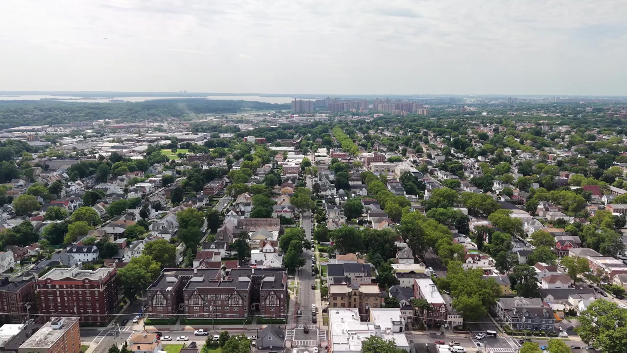 Droneperspektiv, der flyver fremad over en beboelsesgade i en udstrakt amerikansk forstad, med en blanding af lejlighedsbygninger, parcelhuse og frodigt grønne træer på en delvist overskyet dag.