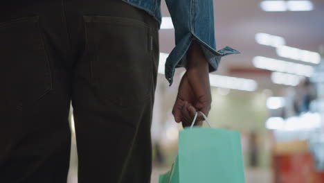 man wearing denim jacket holding green paper shopping bag while walking through modern brightly lit mall corridor with blurred displays and reflective metal rails conveying urban retail lifestyle mood