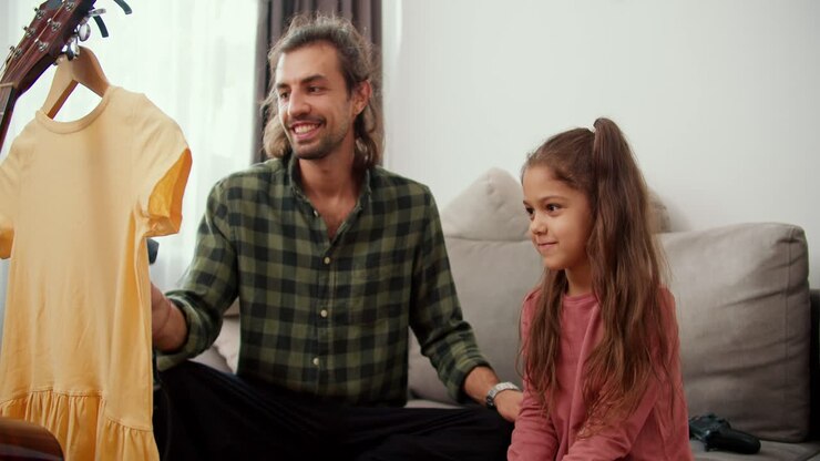 A brunette man with long hair wearing a green plaid shirt together with his brunette daughter, a little girl in a pink jacket steaming a yellow T-shirt using a special steamer while sitting on a gray sofa in a modern apartment