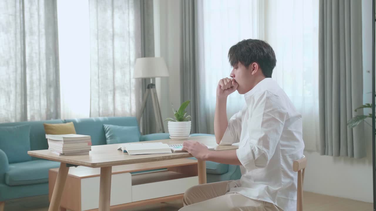 Side View Of Asian Man Student Being Sleepy While Writing In The Notebook On The Table At Home