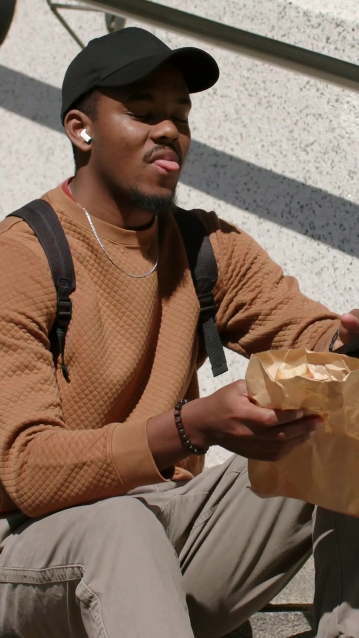Vertical video: Raising brown bag, man in black cap checking phone sitting on skateboard, eating