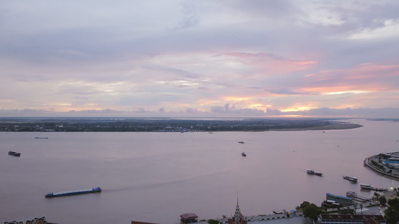 Aerial video of Phnom Penh captures barges and boats drifting along the wide river at sunrise, with soft pastel skies stretching across the horizon