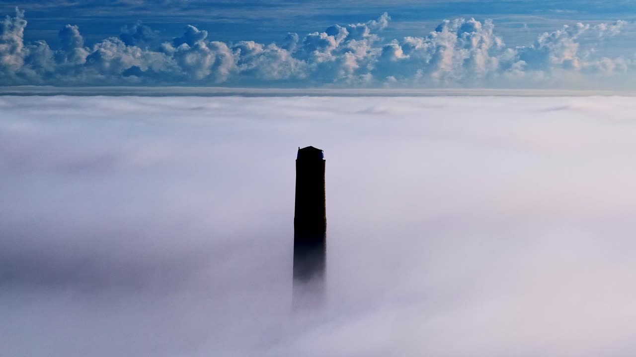 Aerial orbit of dark tall smokestack piercing through thick clouds under a blue sky