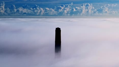aerial orbit of dark tall smokestack piercing through thick clouds under a blue sky
