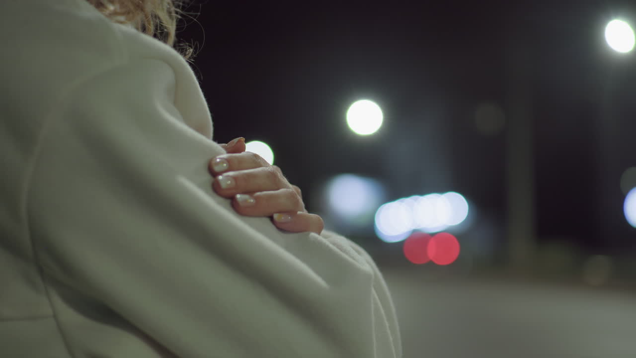 Side view of woman standing outdoors at night with hands folded over chest, showcasing polished nails and soft white coat, background glows with bokeh city lights and blurred motion of passing cars