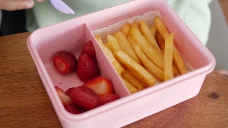 niño comiendo fresas y papas fritas de una caja de almuerzo rosa