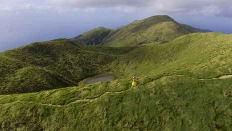 Asiatische-Malaysische-Chinesische-Touristin,-Die-Auf-Einem-Pfad-Am-Rand-Des-Berges-Mit-360-kamera-selfie-stick-Spaziert,-Drohnenansicht-Auf-Pico-Da-Esperança,-Auf-Der-Insel-São-Jorge,-Azoren,-Portugal