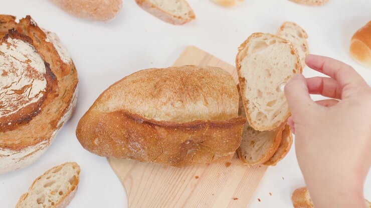 Top-down view of hands slicing crusty sourdough loaf on wooden board with bright lighting.