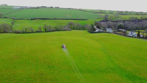 smooth aerial drone view following a red tractor fertilising a greet field with trees from a distance