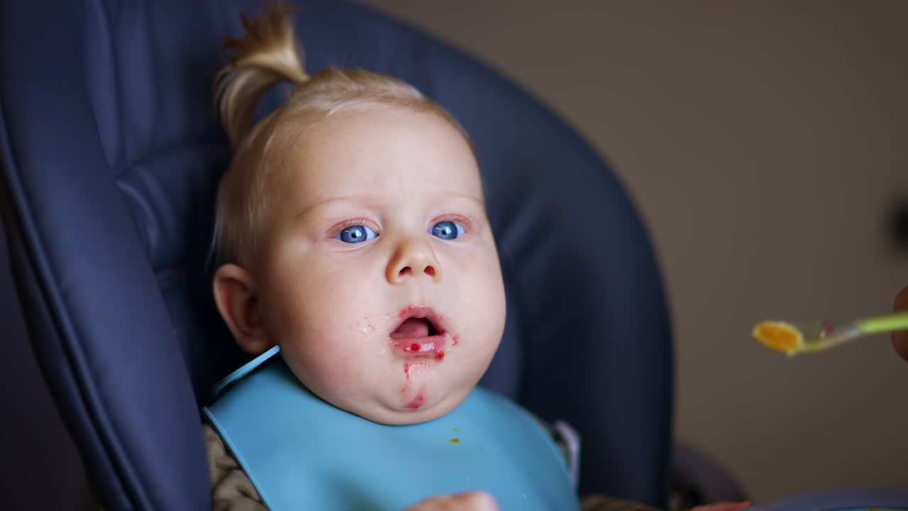 Adorable blue-eyed blond kid is fed from spoon. Lovely baby is focused on camera. Close up.