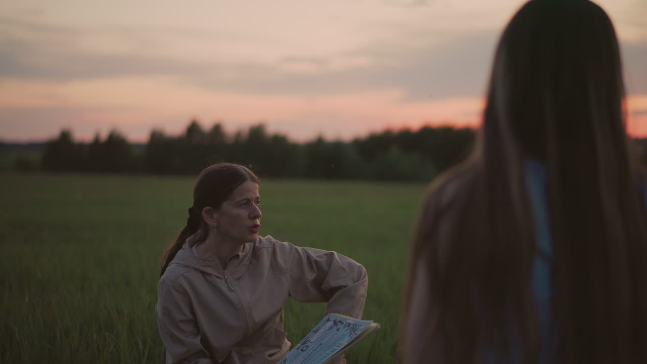 close up back view of woman with long hair seated on grass interacting with another person holding journal at sunset in open field capturing warm storytelling moment under soft dusk light