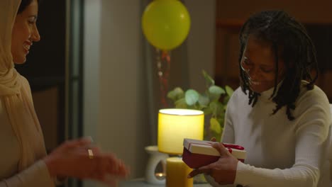 woman giving female friend birthday present at home with party balloons in background 2