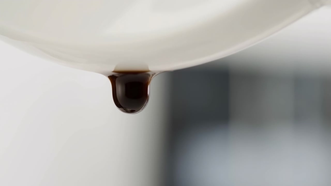 Goccia scura e lucida si allunga e pende dal bordo inferiore di un liscio bordo in ceramica bianco sporco, mostrando una colonna ambrata traslucida vicino al punto di contatto e un bulbo arrotondato marrone scuro sotto con forte riflesso speculare