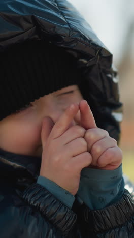 un niño pequeño jugoso sosteniendo sus manos cerca de su cara, cierra ligeramente los ojos, el niño está vestido con una chaqueta negra brillante, con un adulto en una chaqueta marrón cerca, con un fondo borroso
