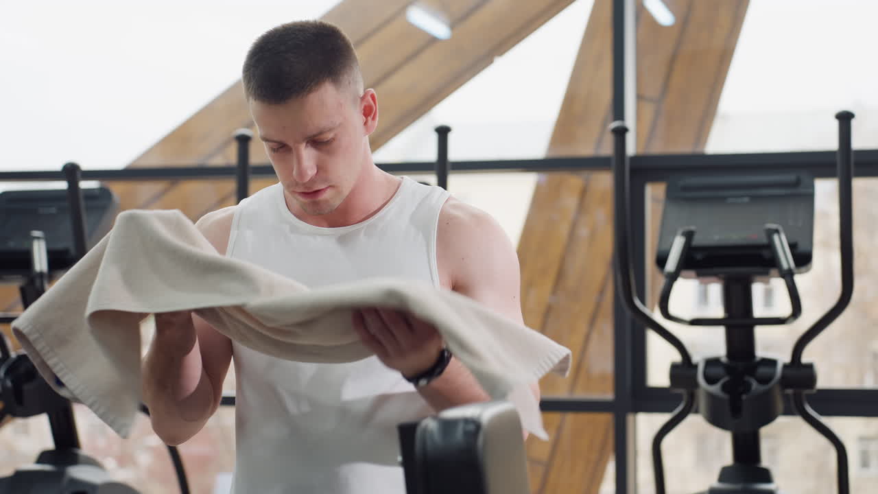 Body built fair guy picks white towel and wipes sweat from intense training session on elliptical machine, looking fatigued as he cools down in bright gym environment with large windows