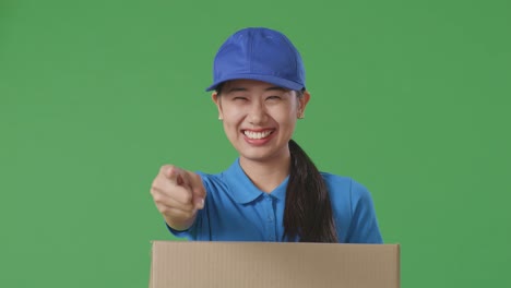 close up of asian female courier in blue uniform smiling and making honest gesture while delivering a carton on green screen background in the studio