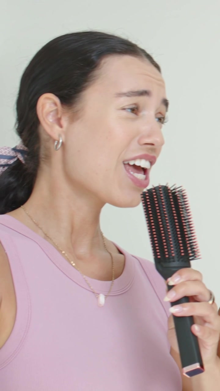 Vertical video: Smiling woman in pink tank singing into paddle brush as friend handing mic at home