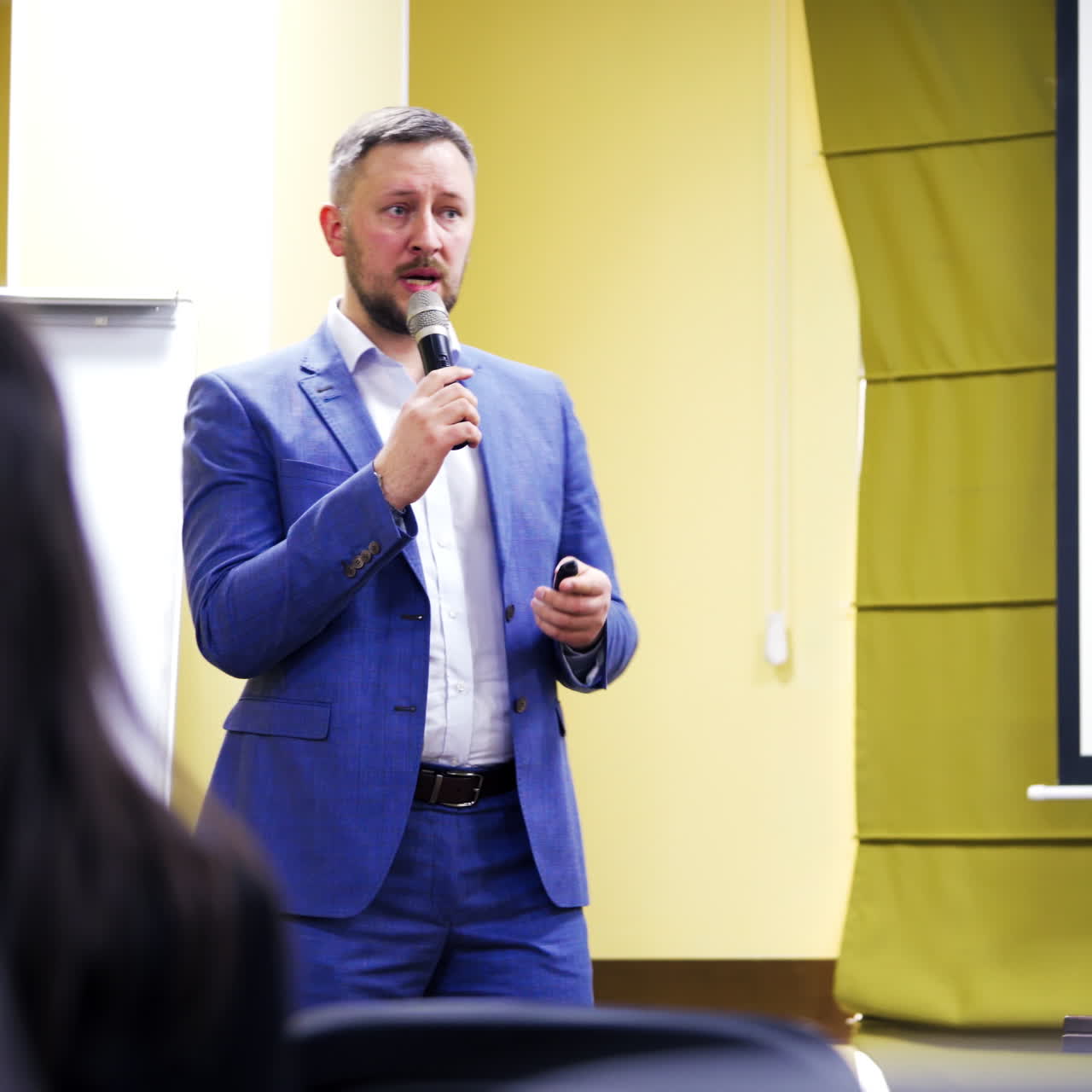 Confident executive manager with microphone speaking at the meeting for the public people. Businessman in blue suit gives advices for audience on business forum.