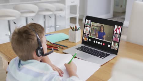 Schoolboy-using-laptop-for-online-lesson-at-home,-with-diverse-teacher-and-class-on-screen