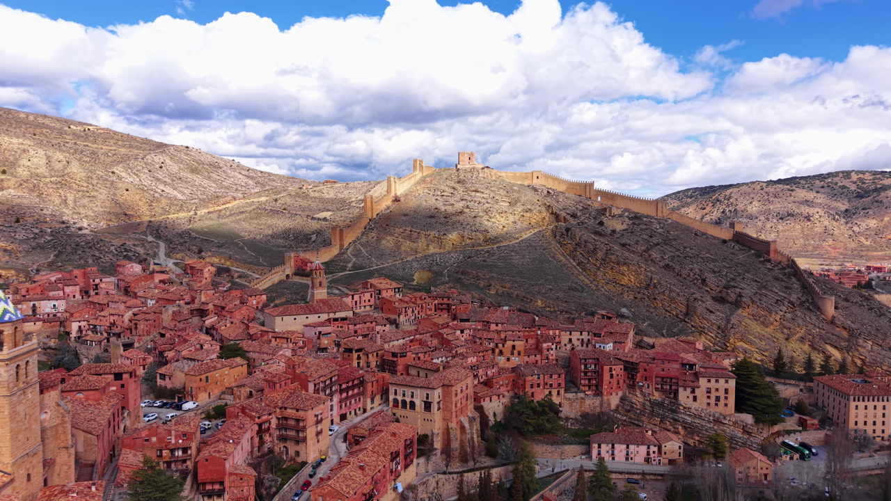 Historic Albarracin city in Aragon, Spain, featuring medieval defensive walls stretching across hillside, terracotta rooftops, church tower and dramatic cloudy sky