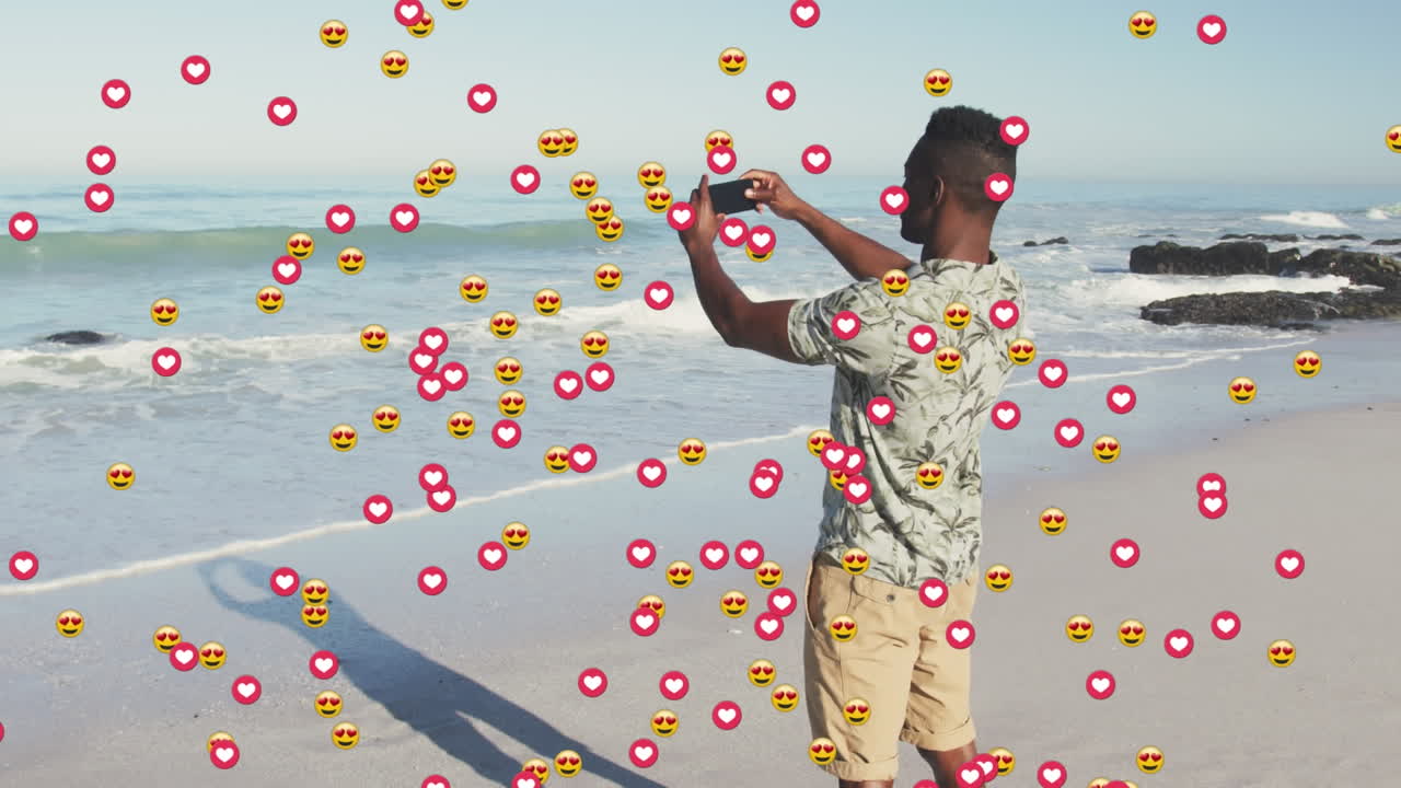 Animation of heart and love emojis over african american man taking smartphone photos on sunny beach