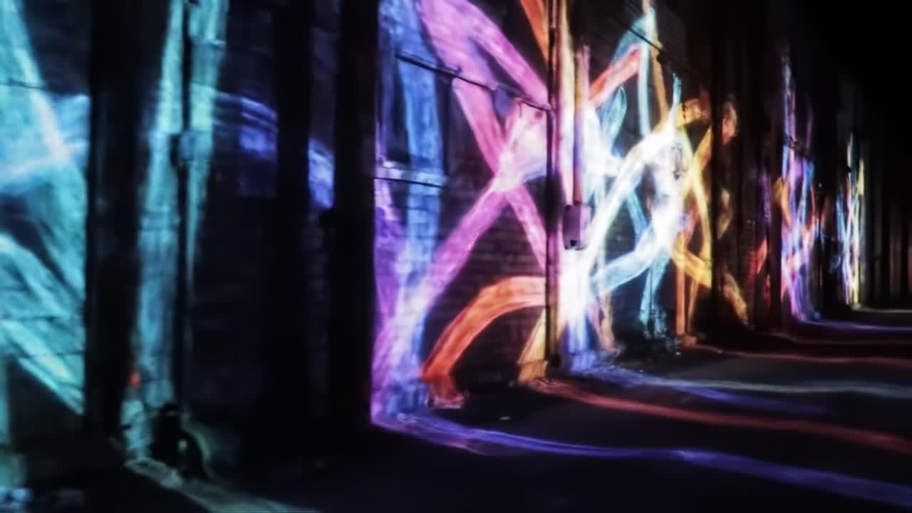Brightly colored light patterns dance on the walls of a city alley, creating a vibrant visual experience. The installation captivates viewers after dark with its dynamic luminosity.