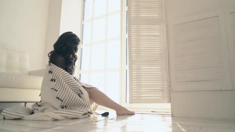 brunette-covered-with-plaid-sits-at-large-window-in-loft