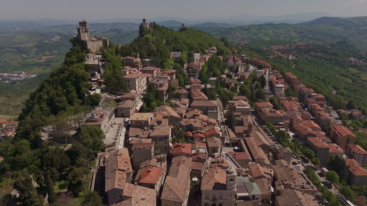 Smooth aerial flyover of San Marino's historic old town on Monte Titano, showcasing La Rocca (Prima Torre), Cesta (Seconda Torre), and Montale. Medieval architecture meets scenic landscapes.