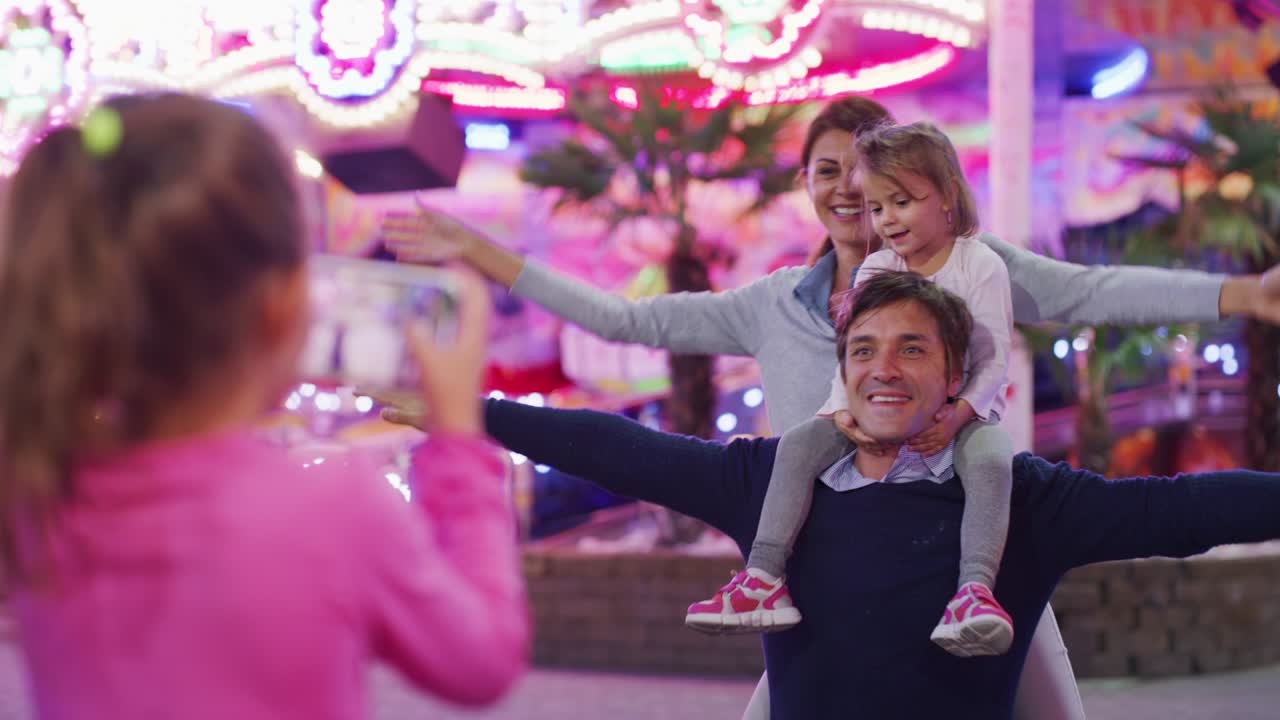una niña feliz se está divirtiendo tomando una foto o un video de su familia mientras disfrutan del tiempo juntos en un parque de atracciones con luces de luna park por la noche.