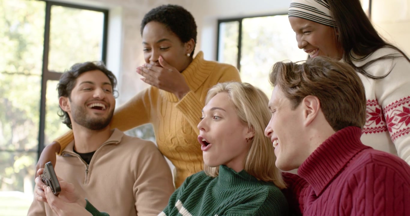Diverse friends at home, man holding phone showing pic, blonde woman in sweater reacting pointing