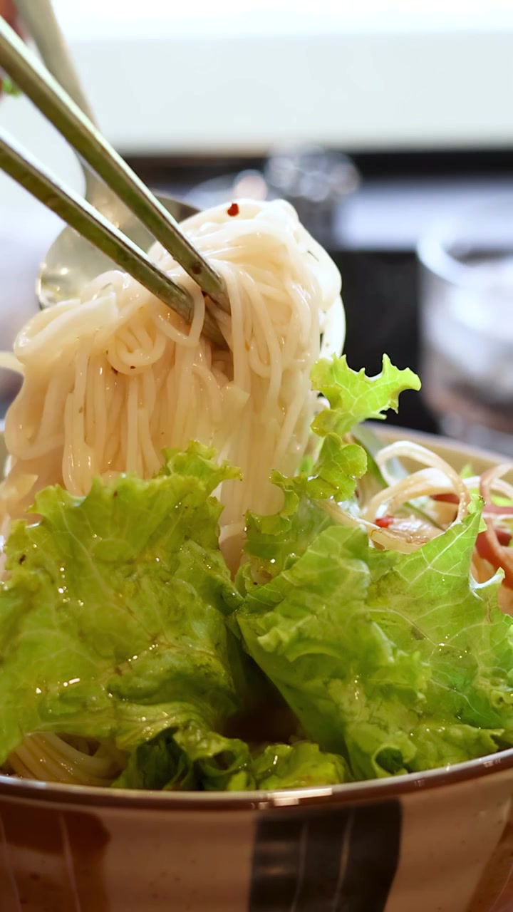 Primo piano di mani che usano bacchette e un cucchiaio per mescolare un'insalata di noodle vietnamita fresca.