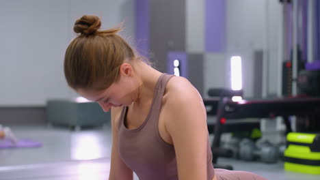 focused woman engaging in deep stretch in a modern gym with bright lighting and mirror reflections, gym equipment and purple yoga mat visible in the background