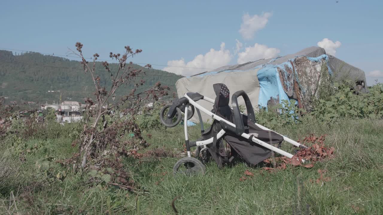 A discarded baby stroller lies in a weedy field in front of a makeshift shack in the Pirita informal settlement, a powerful symbol of childhood hardship and poverty.