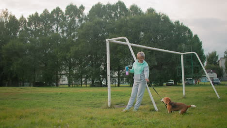 female dog tamer resting against football post while golden retriever energetically plays with leather leash on grassy field during cloudy day outdoor fun bonding moment joyful interaction