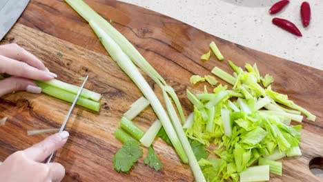 hands use a kitchen knife to slice celery stalks on a wooden cutting board in bright, natural light with a top-down camera angle