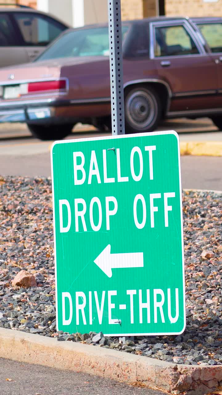 Vertical video of election signage reading “Ballot Drop Off” with arrow points roadside as cars pass, symbolizing democracy, civic duty, inclusivity, freedom, responsibility, safety, and trust