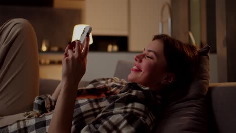 side view of a happy brunette girl in a checkered shirt lies on a gray sofa on a pillow and types something on social networks using a white smartphone rejoices and has fun in a modern apartment in the evening