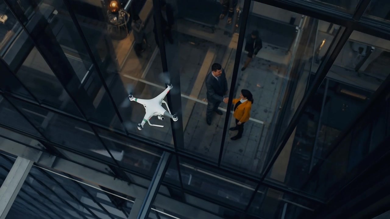 Drone pairando, fazendo com que um homem e uma mulher com casaco mostarda troquem um aperto de mão de negócios em um átrio de vidro.
