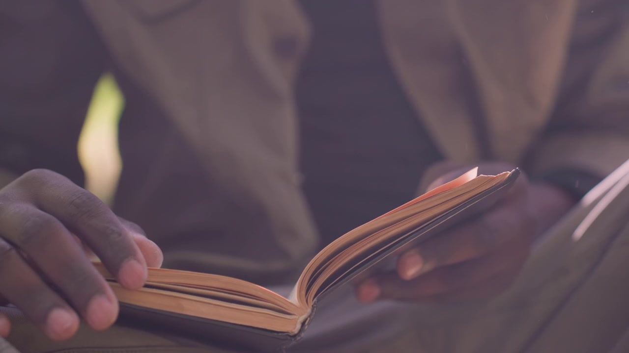 Primo piano di una persona che legge un libro aperto con le mani visibili, concentrandosi sull'interazione tra dita e pagine. La scena mostra un'illuminazione soffusa e costante che evidenzia i dettagli della texture della carta.