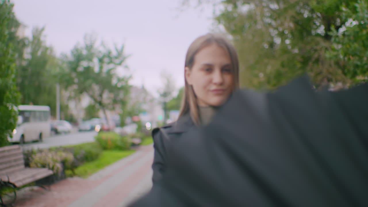 Pretty girl spinning a large black umbrella on city sidewalk with bench cars and trees in blurred background during cloudy day representing urban lifestyle outdoor protection seasonal weather