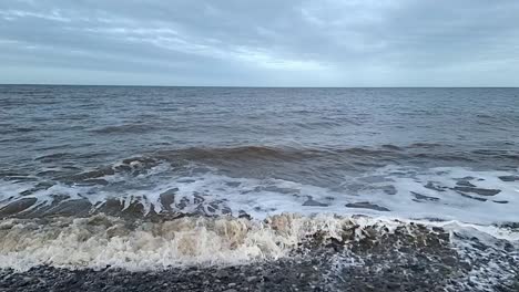 dirty slow motion tide waves crashing onto stormy pebble waterfront looking out across horizon