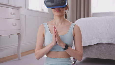 tilting shot of woman sitting on yoga mat wearing virtual reality headset in bedroom at home taking part in virtual class - shot in slow motion