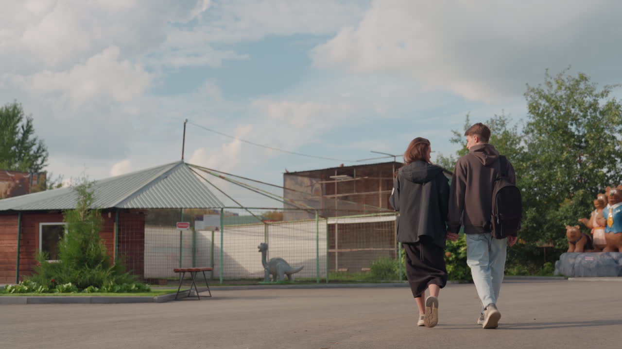 To hvite tenåringer går forbi en paviljong under en skyet sommerhimmel, et kjettinggjerde og rustne boder rammer inn scenen, lyslenker henger over, ryggsekker og hettegensere, sakte reflekterende spasertur, forstad